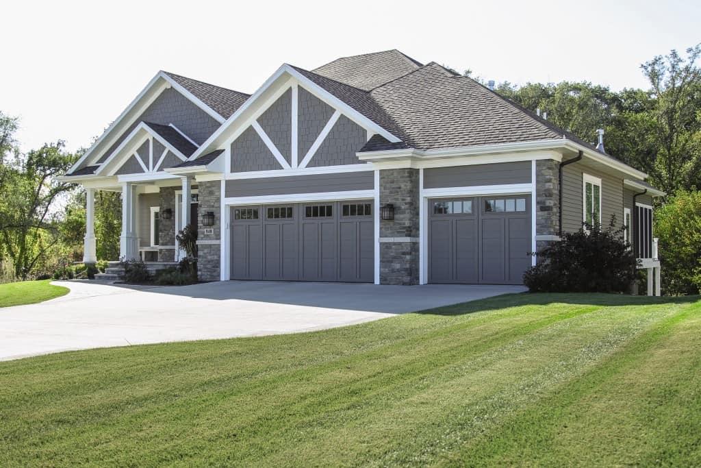 A large, modern suburban home is photographed from a low angle on a sunny day. The house features gray siding, stone veneer accents, and a prominent front area with two double garage doors in a matching gray color. The roof is shingled, and the architectural details include white trim and decorative trusses in the gables. A wide concrete driveway leads up to the garages, and a neatly maintained green lawn slopes in the foreground.